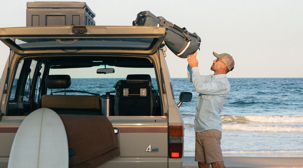 A man on the beach with his 4 wheel drive, surf board taking his North Storm 60 litre duffel bag off the roof. 