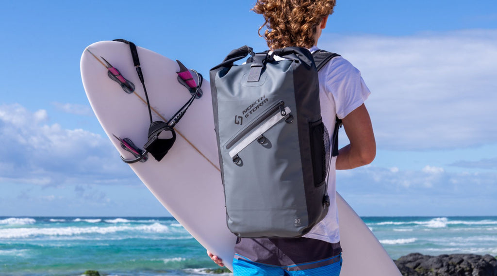 A young man walking along a beach holding a surfboard and wearing a North Storm 30 Litre Backpack.