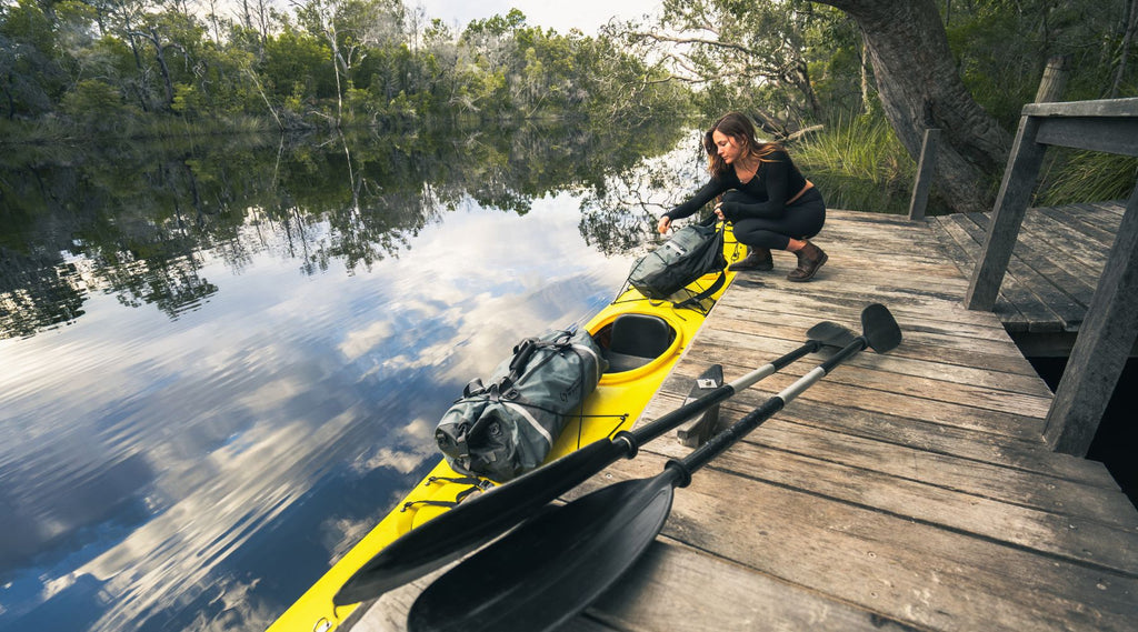 A lady at a wooden jetty on a river placing a North Storm waterproof backpack onto a kayak. The kayak also has a North Storm 60 litre Duffel bag on the back.