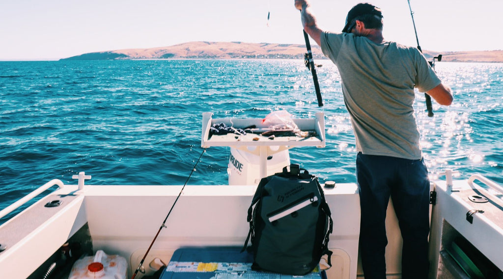 A man fishing on a boat out at sea. He is placing a rod back into a rod holder. There is a North Storm waterproof backpack on the deck keeping his gear dry. 