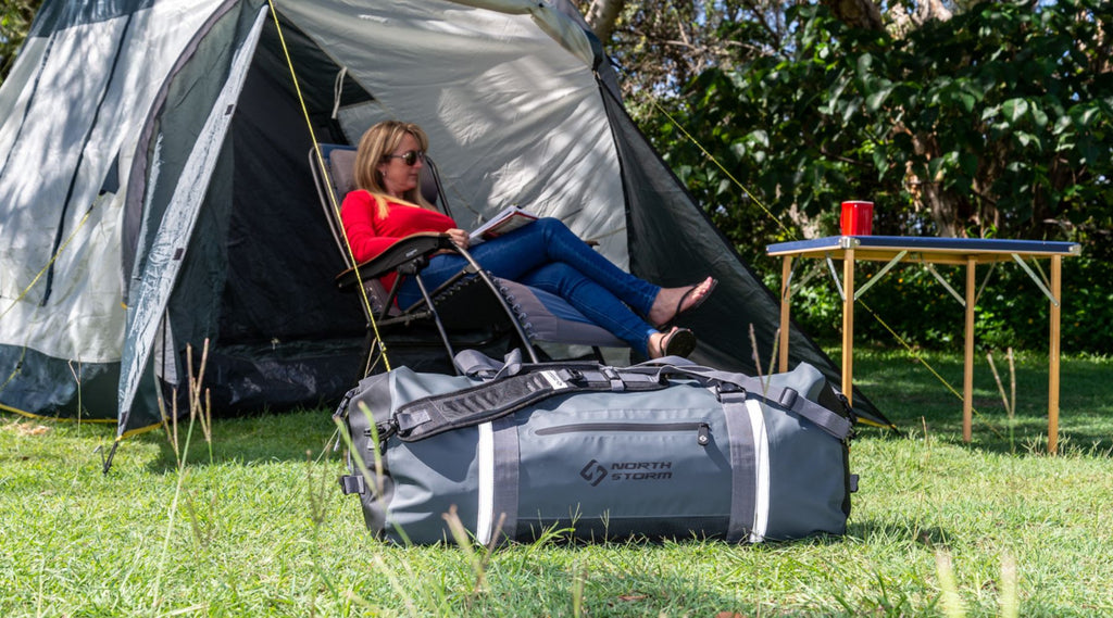 A lady sitting relaxing in a fold out chair at a campsite. She is happily reading a magazine. There is also three waterproof bags in front of her on the grass at the campsite. 