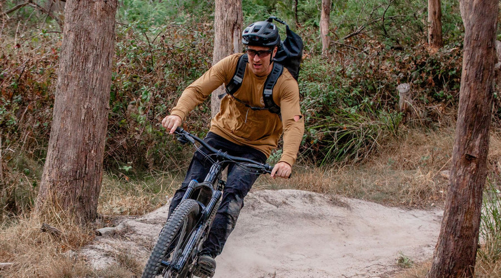 A man smiling and riding a mountain bike on a dirt track through the bush. He is wearing a helmet and a North Storm 30 L waterproof backpack.