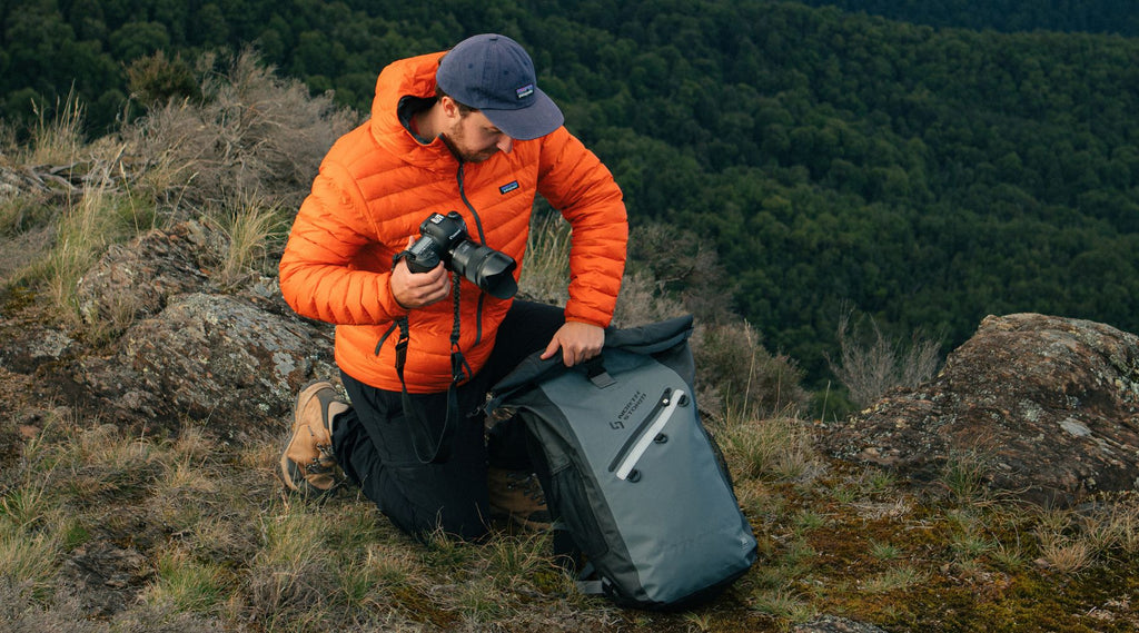 A man wearing a thick warm jacket, kneeling down and holding a camera in the outdoors. He is about to open his North Storm 30 Litre waterproof backpack.