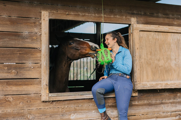 Caitlin sitting with a brown horse, smiling while holding a green Jolly Hay Ball filled with hay and carrots