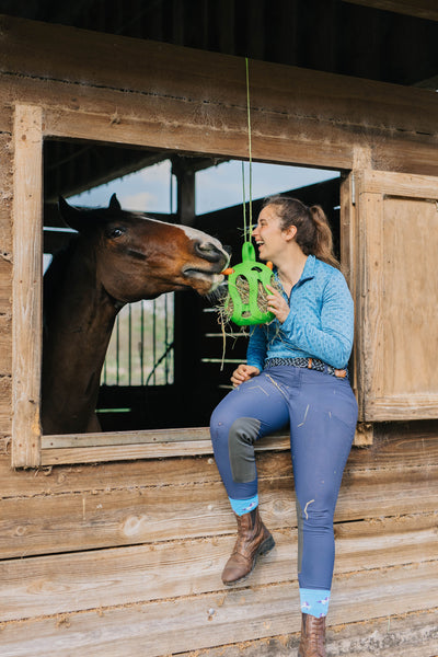 Caitlin sitting with a brown horse, smiling and holding up a Jolly Hay Ball stuffed with hay and carrots