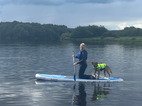 dog standing on back of paddleboard on water whilst owner kneels in-front paddling