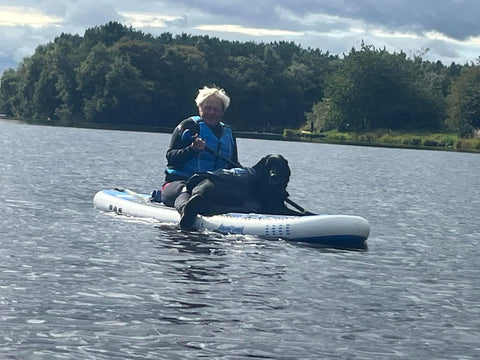 black Labrador sitting on front of paddleboard with owner sitting on same board