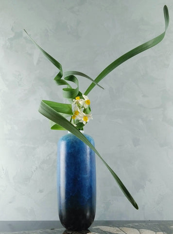 Nageire-style Ikebana tall blue ceramic vase with green leaves and white and yellow flowers