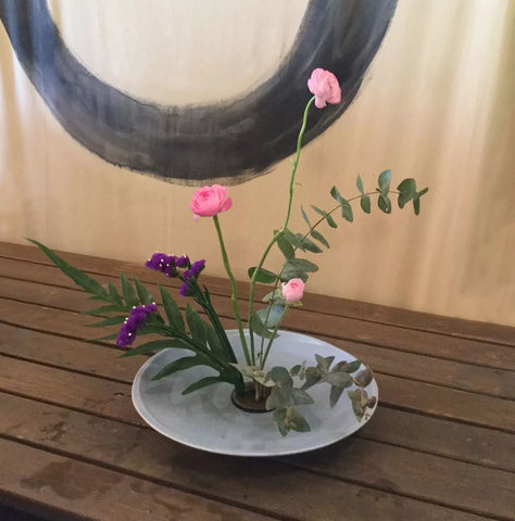 Ikebana estilo Moribana con flores pequeñas rosas y gladiolos violetas y amarillas con hojas de diferentes plantas de tonalidades verdes