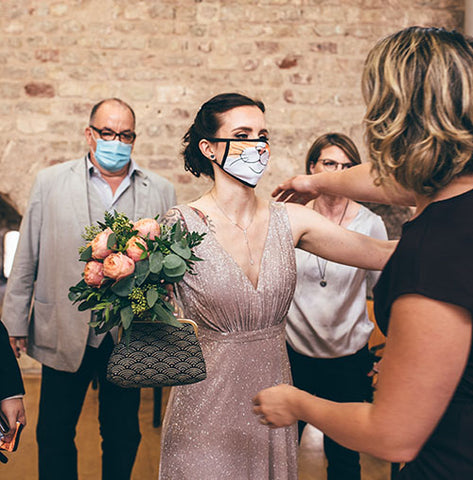 A lovely young bride holding a bouquet and wearing a cat-inspired face covering embraces a member of her wedding party.