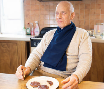 Older man in a kitchen eating while wearing the protective scarf bib in steel blue.