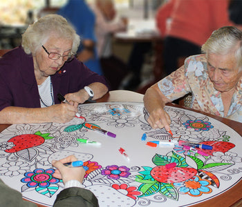 A group of older ladies coloring the giant accessible art dry-erase board together, that takes up all the table.