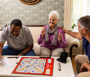 An elderly woman, with two middle-aged men, smiling while playing the inclusive and adapted Scrabble Bingo.