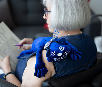 Older lady reading with the Manimo weighted plush blue cat on her shoulder.