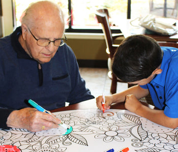 A grandpa and his grandson coloring a giant art erase-board together at a table.