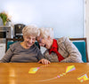 Two elderly women, sitting and smiling around a table, playing a Tovertafel game with kites.