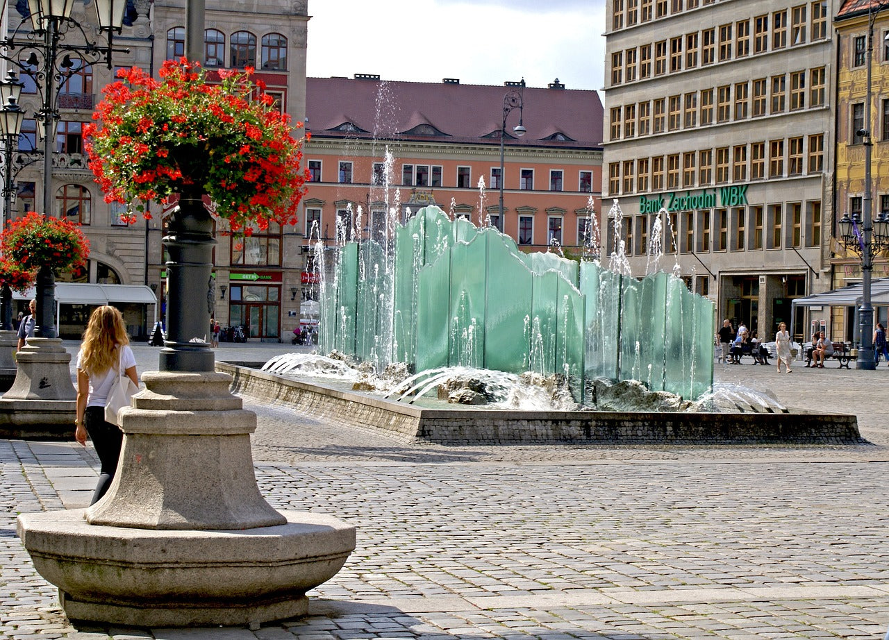 A woman near a large, glass-paneled fountain in a European city square.