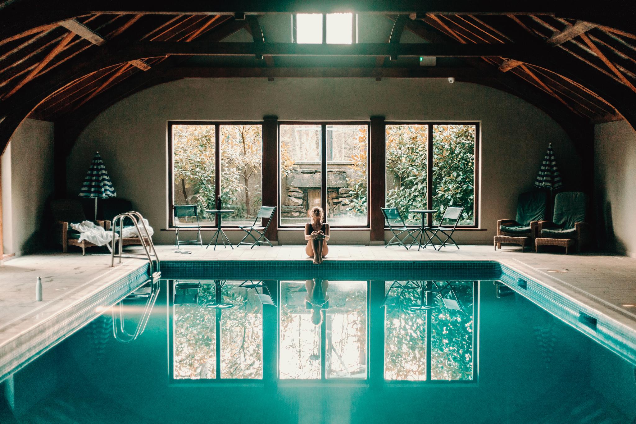 Person sitting by an indoor swimming pool with large windows overlooking trees.