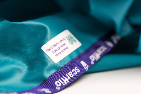 This image shows a close up of the care label of a Wild Teal scarffio® which has contrasting purple branded edging. The care label has a Teal Dragon on it, and states Wash at 30°C, Do not Tumble Dry, Made in Wales. 