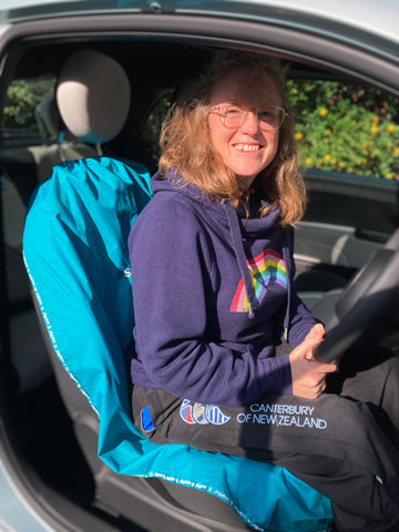 This image shows a smiling lady sitting in the drivers seat of a car, on a vibrant Wild Teal scarffio®. the lady is wearing a navy hooded jumper with a huge rainbow on it, and bright embroidered tracksuit trousers. Demonstrating how having a scarffio® is the bright way to protect your car seats. 