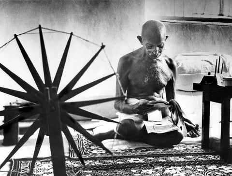Foto in bianco e nero di M. Burke-White: Gandhi legge vicino un filatoio in primo piano, simbolo della lotta per l'indipendenza.