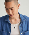 A man wearing a blue shirt showcases the Victory: Winged Coin Charm Pendant, featuring a silver medallion inspired by the Greek goddess Nike.