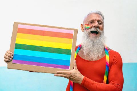 Older gay man in red shirt and rainbow suspenders holding Gilbert Baker Rainbow Pride Flag on cardboard