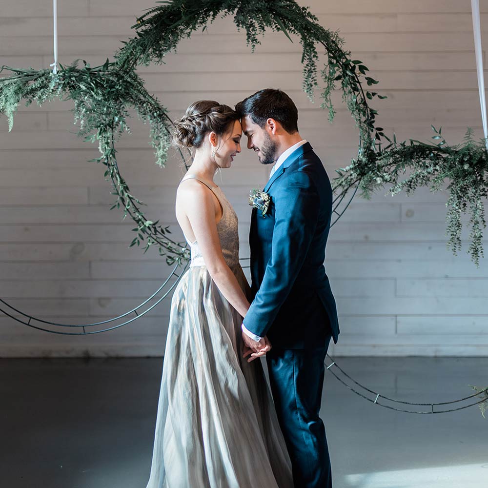 Silver grey wedding dress and navy suits on a couple for wedding. Standing in front of Greenery wreaths