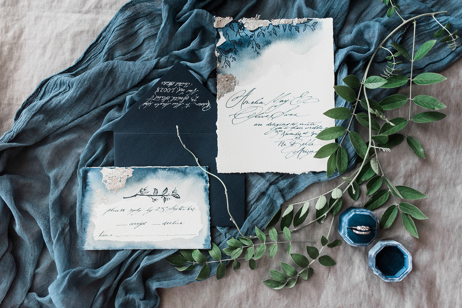 silver foil wedding invitations with elegant relaxed calligraphy for a personal touch. Wedding rings and greenery in a flat lay