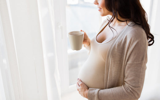 Schwangere Frau steht am Fenster und trinkt Kaffee