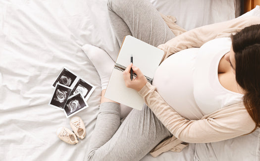 Schwangere Frau sieht Ultraschallbilder an und schreibt Babynamen in ein Notizbuch. 