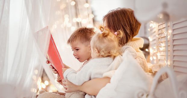Eine Mutter sitzt mit ihren zwei Kindern auf dem Schoß in einem stimmungsvoll beleuchteten Zimmer. Sie halten ein rotes Heft in den Händen.