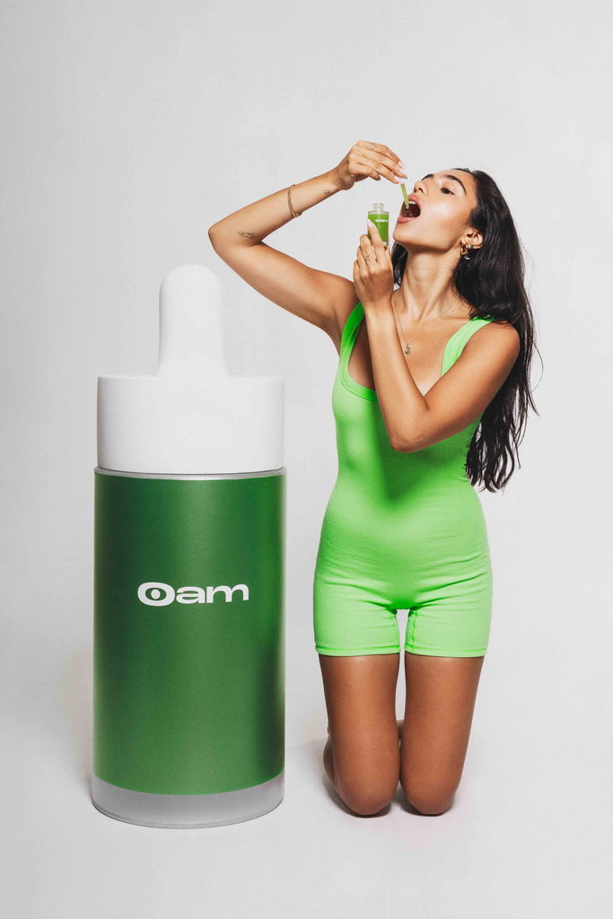 Roxy in a green swimsuit, kneeling next to a life-size eyeam dropper bottle, dropping some of the serum onto her tongue.