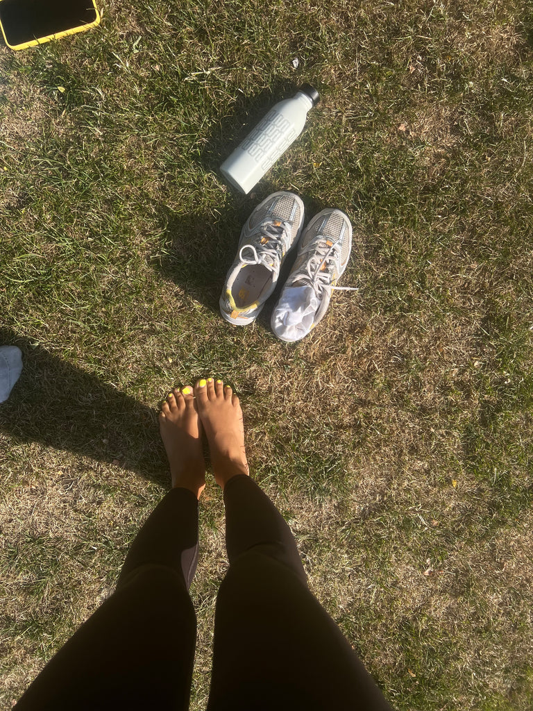 Roxy's legs in exercise gear. She is standing on the grass on a sunny day with her toenails painted lime green. There is a water bottle on the ground, along with her phone and trainers.