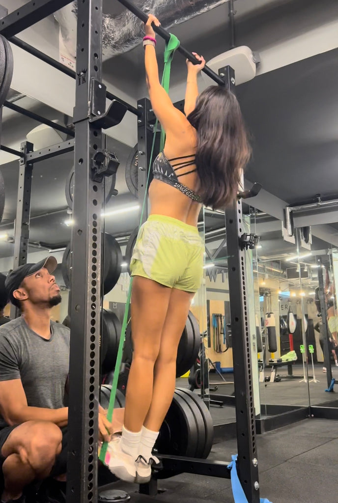 Roxy in the gym on the pull up bar. There is a male fitness coach spotting her.