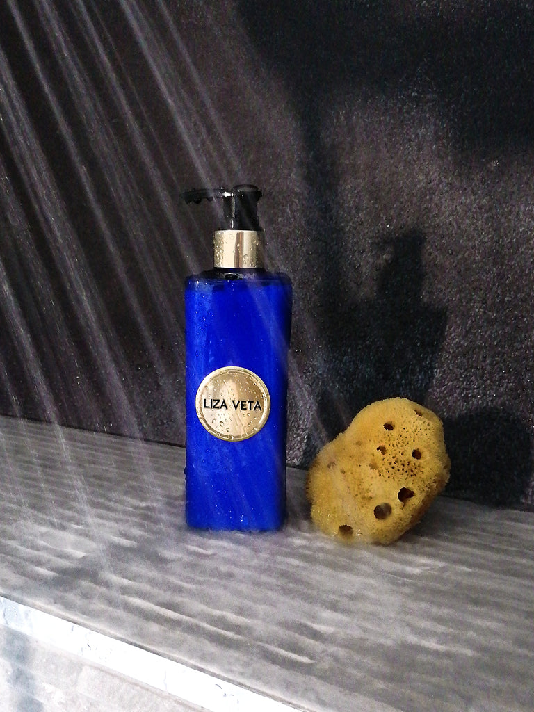This is a Jasmine natural hand & body lotion in blue packaging. In the background, water droplets can be seen falling onto grey tiles. A natural body sponge is also visible on the shelf next to the lotion. The image is related to personal hygiene and shower products and how to take care of yourself during spring.