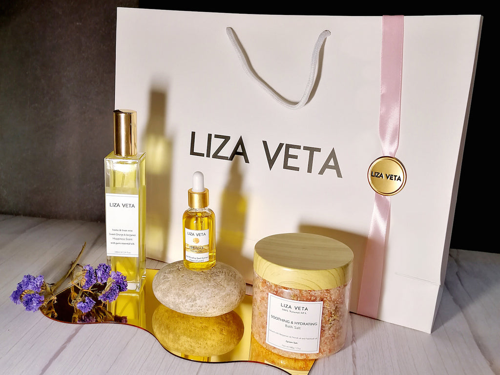 This is a gift set that includes room and linen spray in a glass square bottle, natural face oil and bath salts in a transparent jar. There is a purple flower next to it. This image is related to how to choose a perfect gift set.