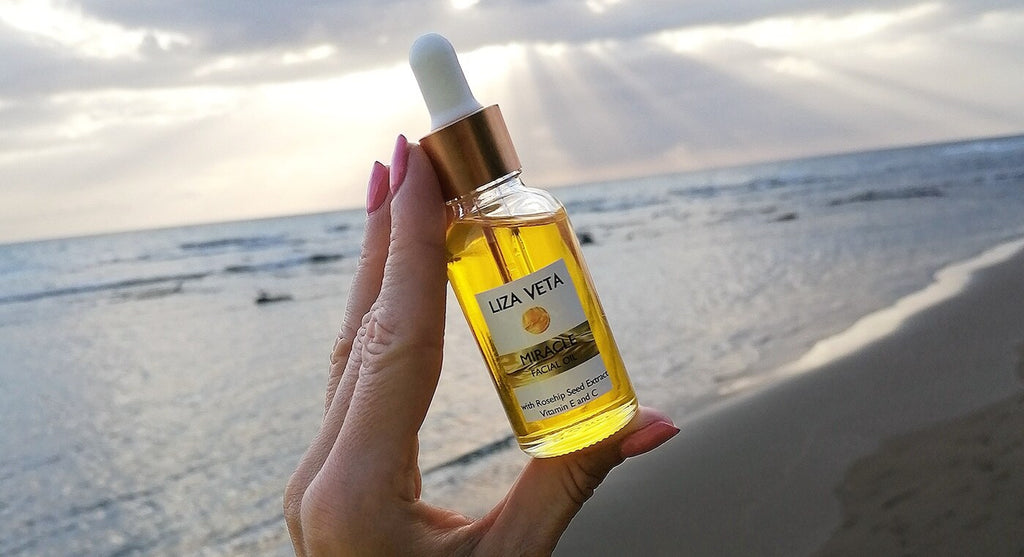 Hand holding glass bottle of facial oil with ocean sunset in the background.