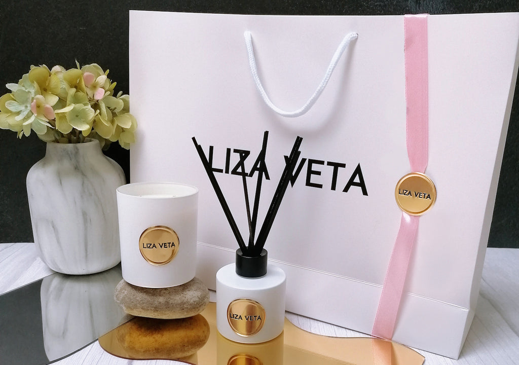 This is a luxurious gift set that includes an Aromatherapy Reed Diffuser and a natural Candle, both housed in a stunning white glass container. There is a branded gift bag with gold logo and pink ribbon. This image is related to how to choose perfect gifts.