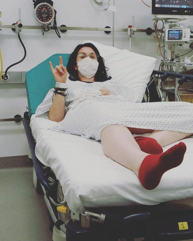 Sarah liggend in een ziekenhuisbed. Ze heeft donker, krullend haar. Ze draagt ziekenhuis kleding, een wit mondkapje, een bril en rode sokken. Met haar hand maakt ze het rock-symbool.