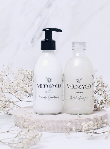 Two Moo & Yoo products (a Miracle Conditioner and a Miracle Shampoo) are sitting on a circular ceramic dish. There is a marble backdrop and the products are surrounded by white blossom.