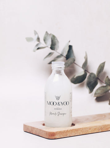 A bottle of Moo & Yoo Miracle Shampoo is sitting on a wooden board with a cream backdrop. There is a sprig of eucalyptus against the backdrop.