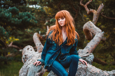 A model with red long flowing hair styled in textured loose waves. Her face is framed with a straight fringe. She is sitting on a tree with a woodland background looking down towards the ground.