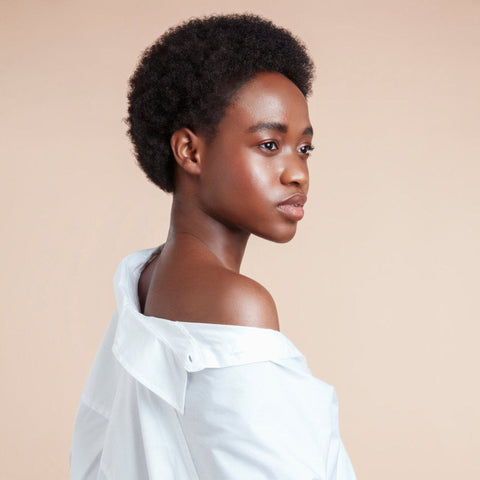 Female model with black natural textured hair hydrated and styled from using Moo and Yoo Miracle Shampoo, Mask, Miracle Milk and Miracle Curl Cream. She is wearing an off the shoulder white shirt and is facing the back wall looking over her shoulder to the side showing her profile