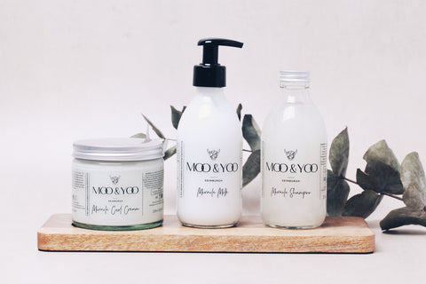 Three Moo & Yoo products lined up on a wooden plank with a piece of eucalyptus behind them. The products are from left to right a glass jar of our miracle curl cream, a glass bottle of our miracle milk and our miracle shampoo.