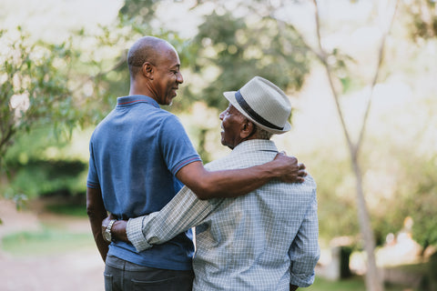 Elderly father with his arms around his sons shoulder.  Looking at one another as they walk in a park.