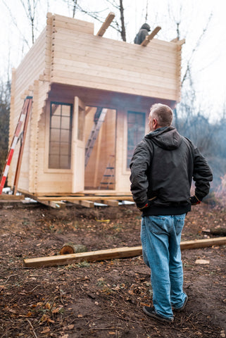 man standing in front of a half finished construction of a wooden tiny house in the woods