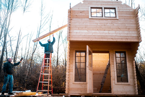 2 men passing a 2x4 wood up a ladder onto a wooden tiny house that they are building in the woods