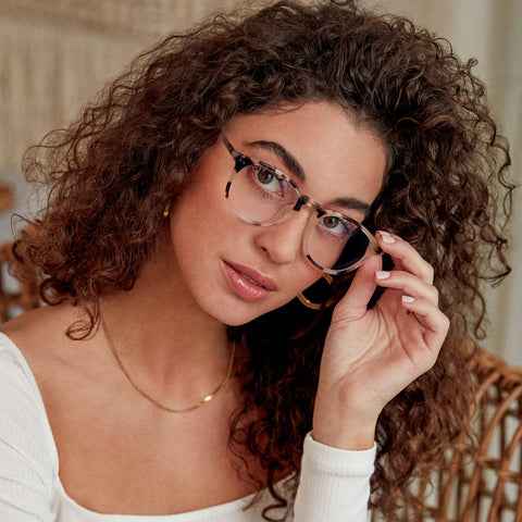 Young lady with long wavy hair on a wicker chair looking into the camera while wearing the Jaxson cream tortoise blue light glasses