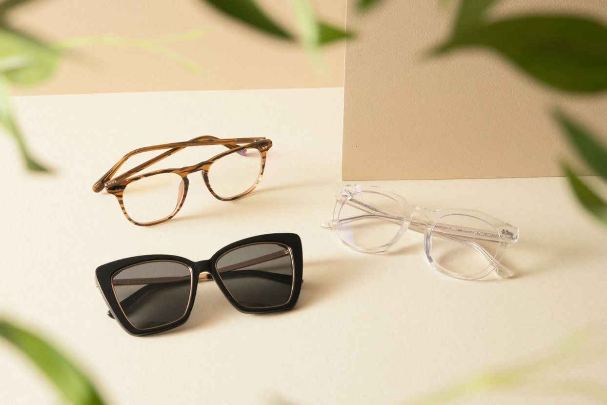 DIFF sunglasses, blue light glasses, and prescription eyeglasses flat-lay on tan surface with green leaves 